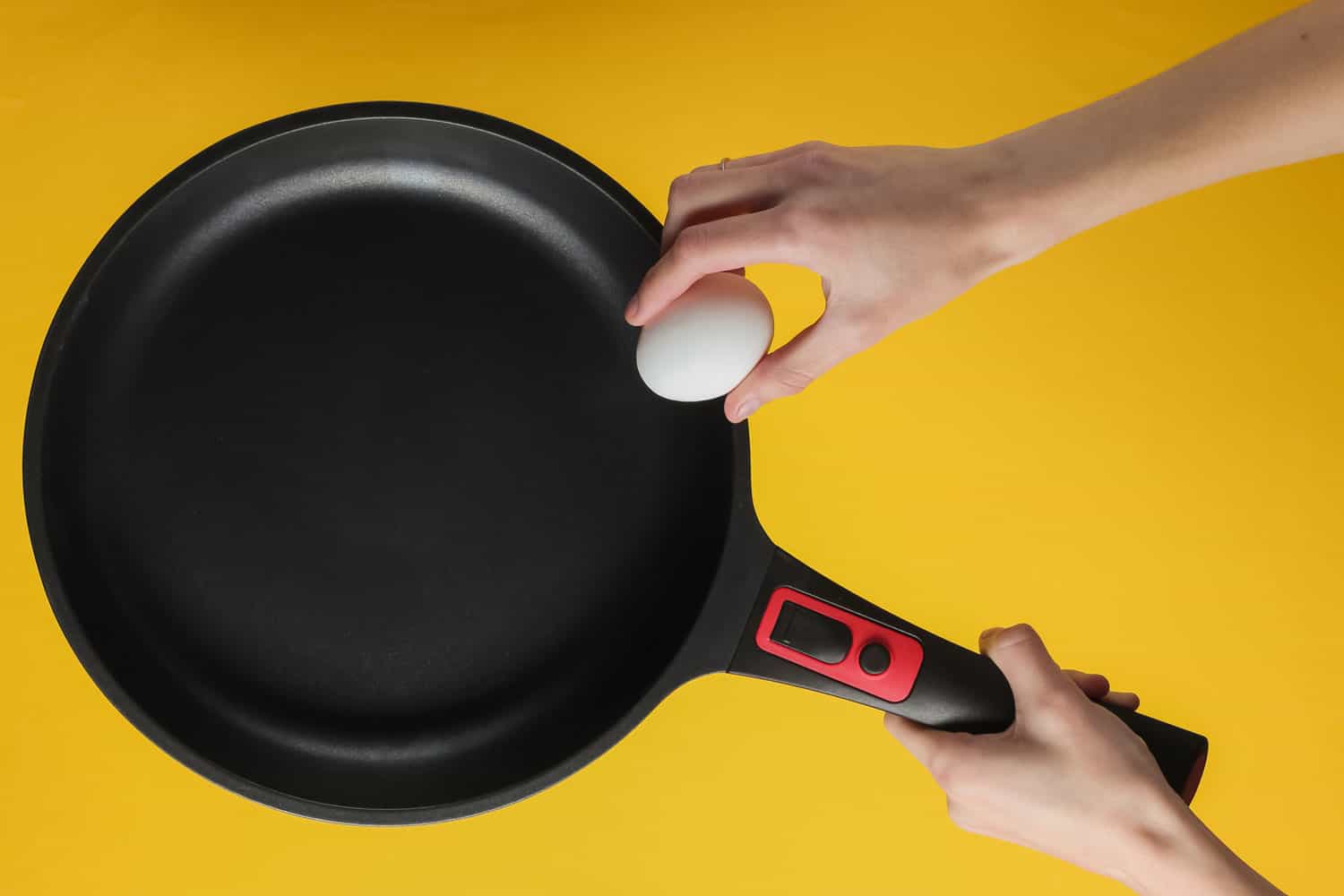 女性的手将鸡蛋煎锅上不粘涂层。烹饪概念。影棚拍摄在黄色背景。”width=