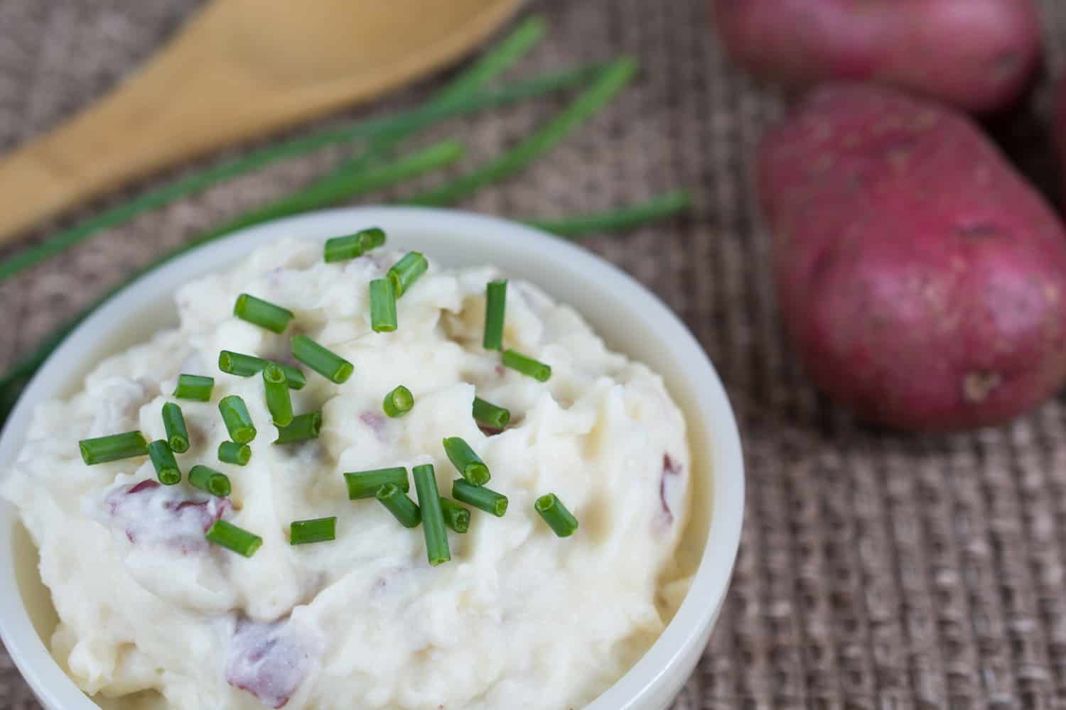 碗自制的土豆泥坐在一个角落里用新鲜切碎的香葱撒在上面。木勺,联合国减少细香葱和新鲜的红土豆位于背景。