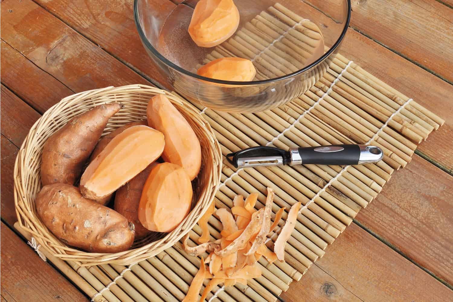 甜土豆在篮子和玻璃碗放在竹餐具垫。削皮器和土豆皮。