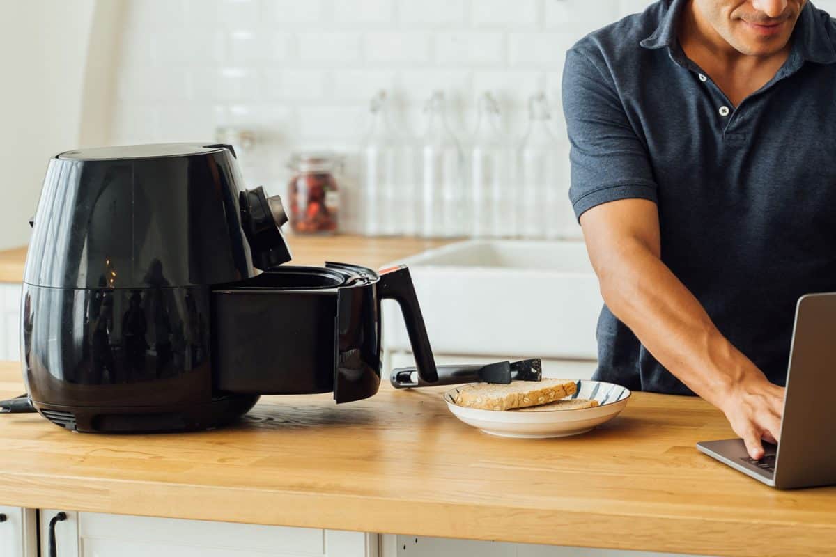 年轻英俊的白人男子看着笔记本电脑上的说明如何使用现代空气炸锅烹饪健康的食物而不使用油在家里的厨房，你可以用空气炸锅代替你的微波炉吗?雷电竞官网网址
