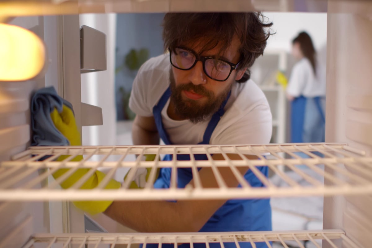 从内部空冰箱的男人戴着橡胶手套和围裙清洁货架上。男性看门人洗肮脏的冰箱用喷雾器和布。房屋清洁服务理念