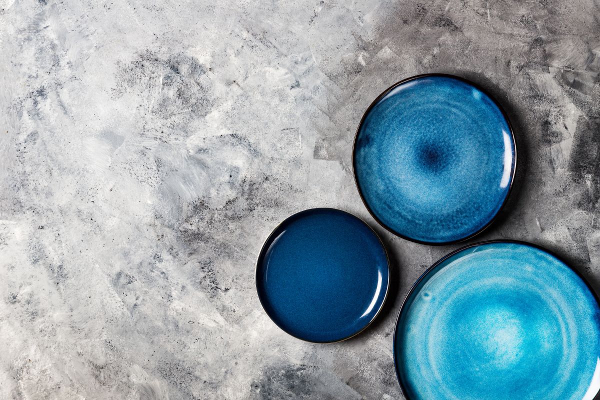 不同的陶瓷空蓝色板在灰色背景，顶视图，复制空间”width=