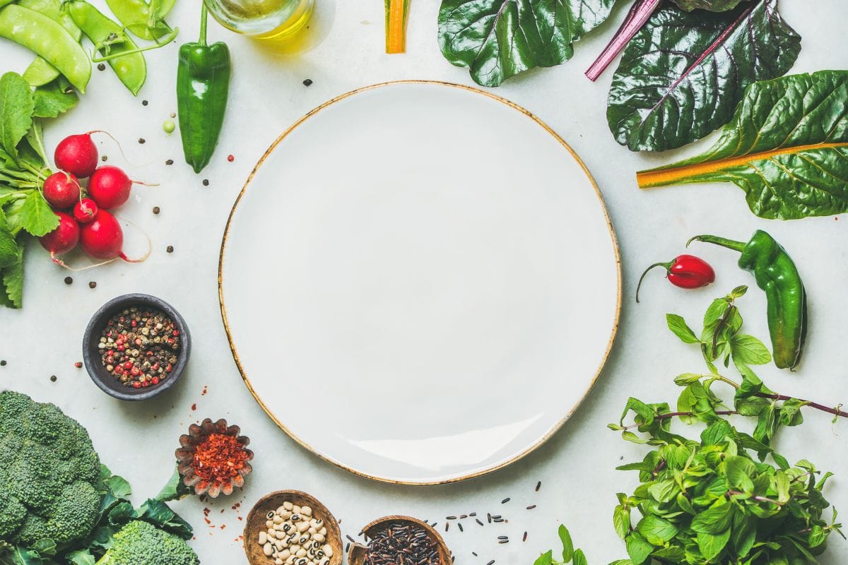 新鲜的生蔬菜，未经加工的蔬菜和谷物放在浅灰色大理石厨房台面上，白色盘子在中央，俯视图，复制空间。雷电竞官网网址健康、清洁饮食、纯素、排毒、节食的饮食理念”width=