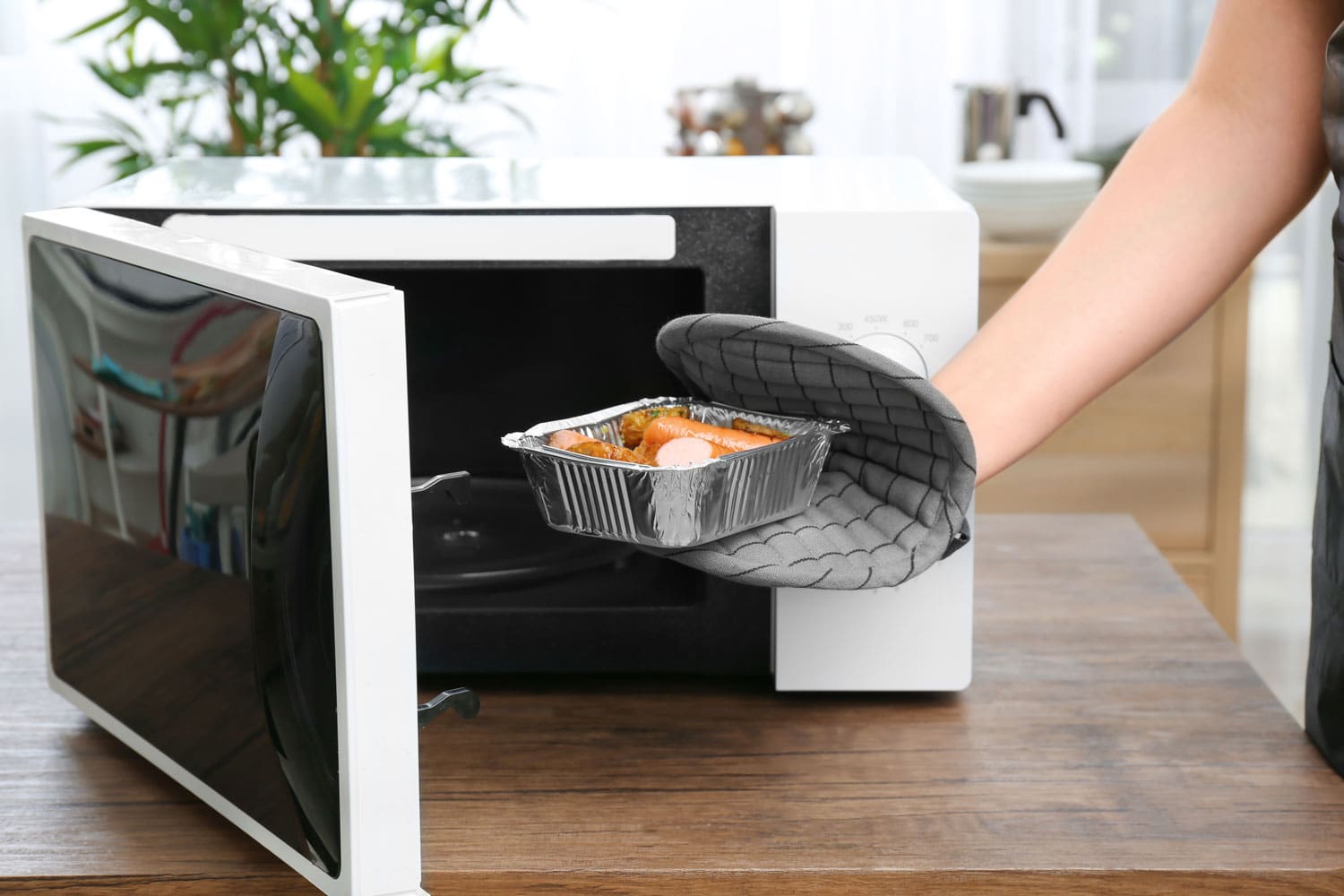 Woman taking foil baking tin with potato and sausages out from microwave in kitchen