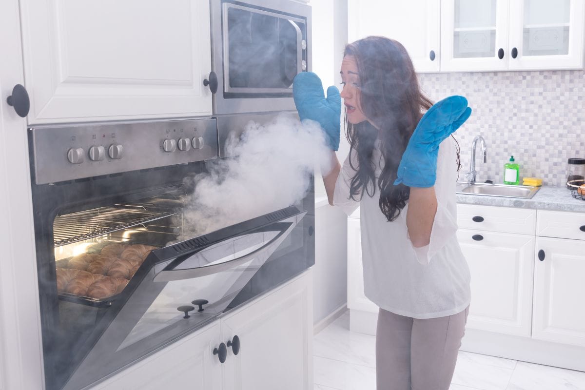 惊讶的女人站在燃烧的烤箱前，厨房周围有烟。雷电竞官网网址-为什么我的烤箱在预热时冒烟?[&如何解决?]］